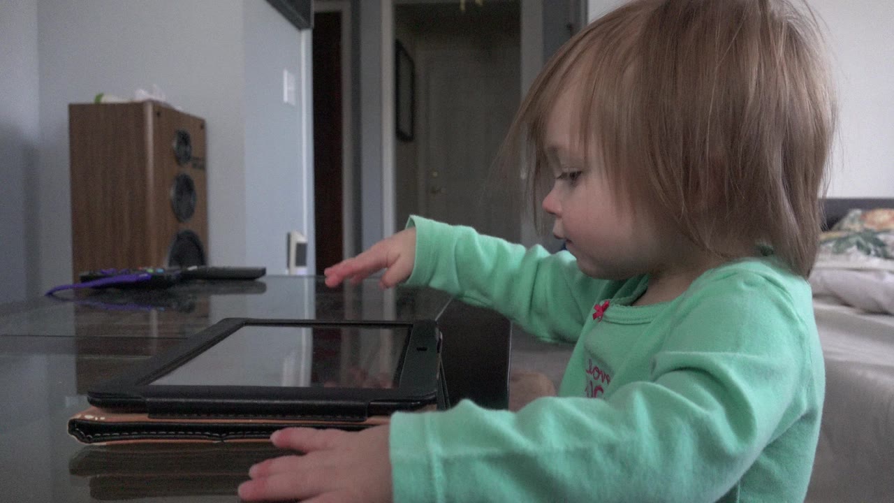 At 0:00 in Westminster, Colorado, a toddler in a mint green top reaches toward a tablet on a glass table while sitting beside a couch in a home interior.