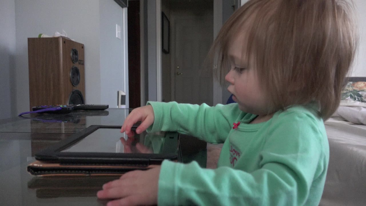 In Westminster, Colorado, a blond young girl in a mint green shirt reaches toward a tablet on a glass table beside a bed and doorway.