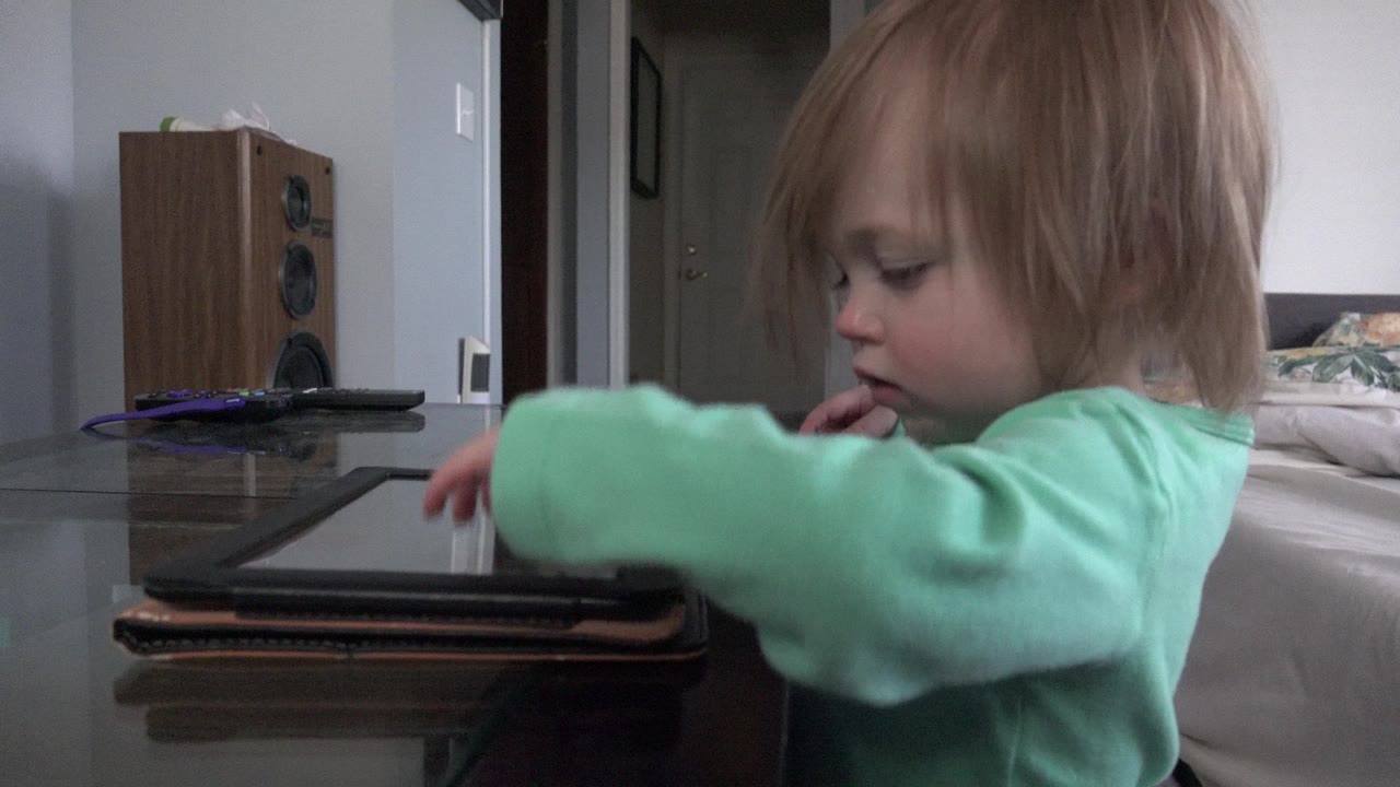 Young child in a green top reaches toward a tablet on a glass table.
