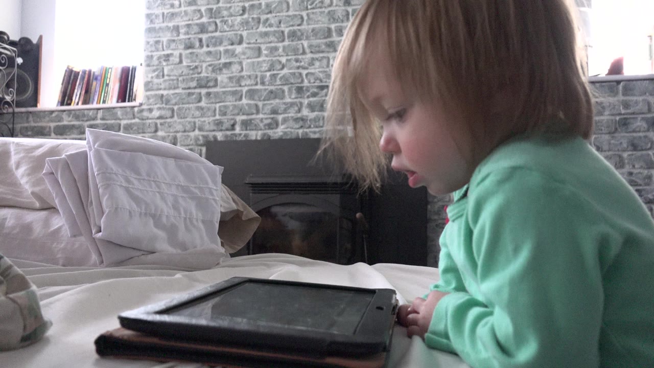 Young child leaning over a tablet on a bed.