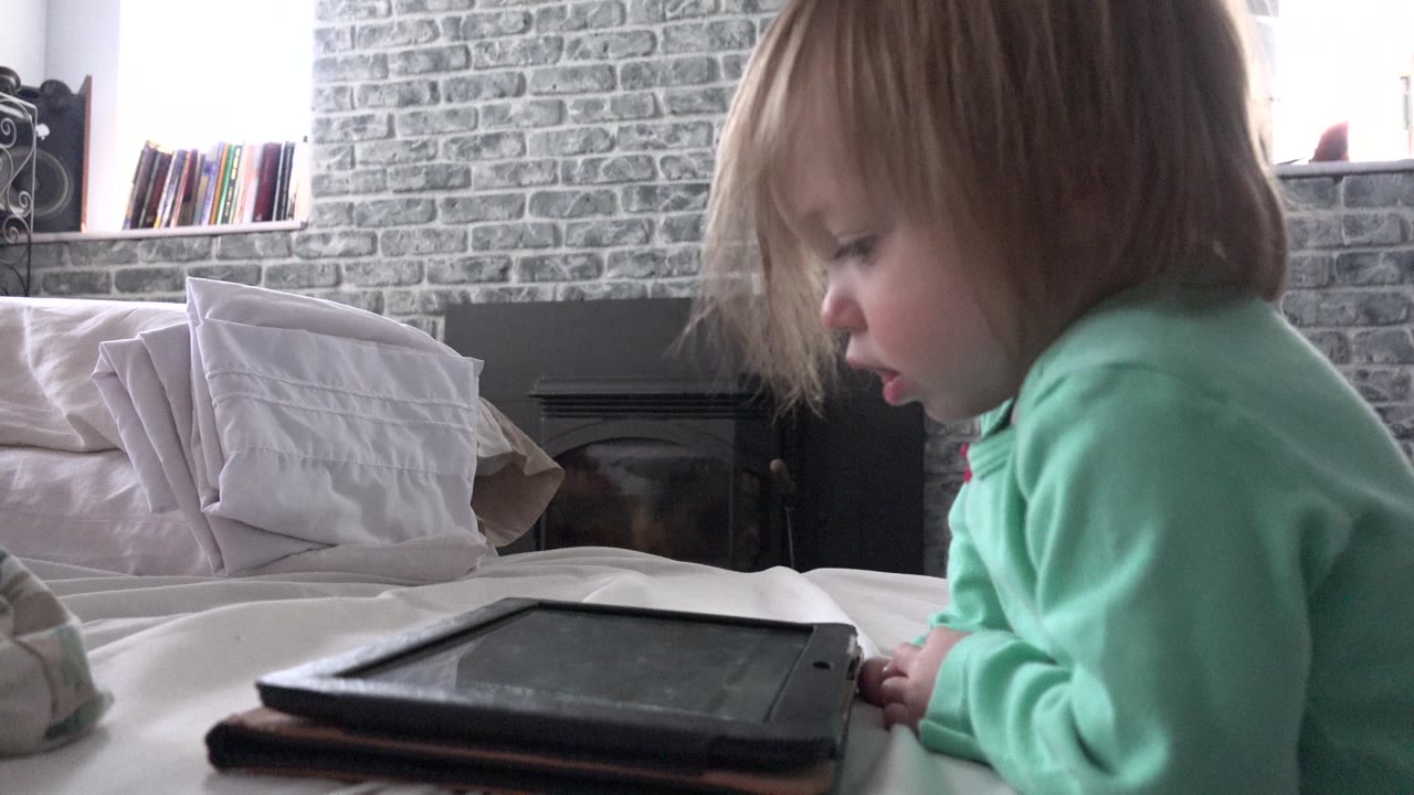 At 0:01, the child remains in side profile, looking down toward the tablet while pale pillows and a dark fireplace sit behind the bed.