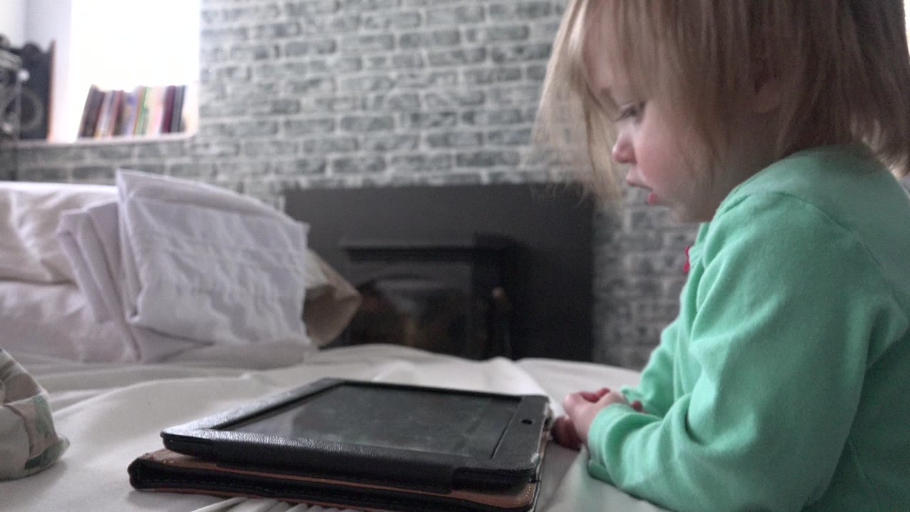 Young child in a green shirt lying on a bed near a tablet.