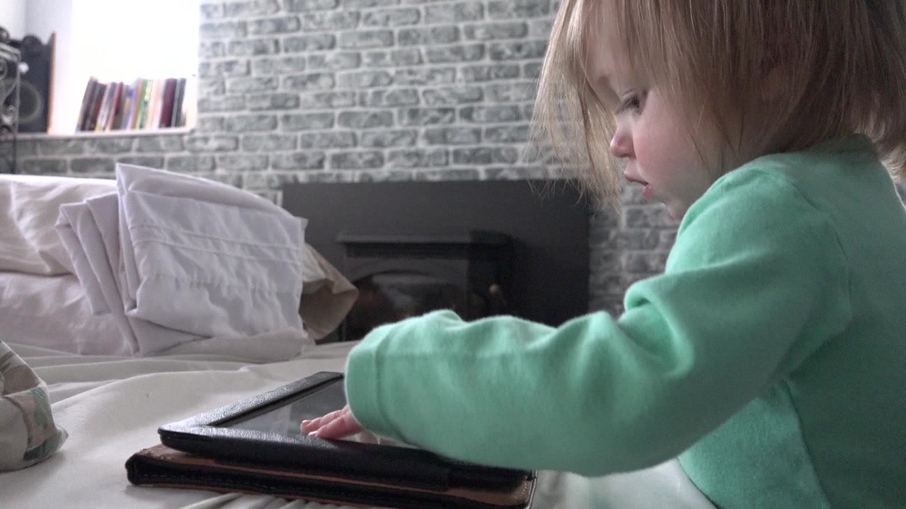A toddler in Westminster, Colorado keeps one hand on the iPad screen while sitting beside pillows on a bed in a softly lit room.