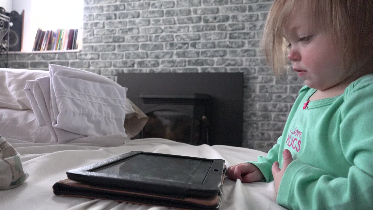 A blonde toddler in a mint green shirt sits at the right side of a bed in Westminster, Colorado, with a black tablet lying in the foreground and pillows behind it.