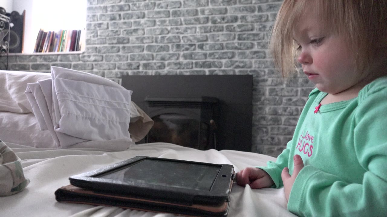 The toddler looks downward beside the black tablet on the white bed, with the gray brick-pattern wall, pillows, fireplace, and bright window visible in the Westminster home interior.