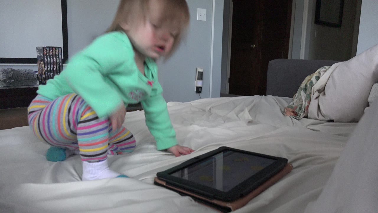 Toddler crouching on a bed near a tablet.