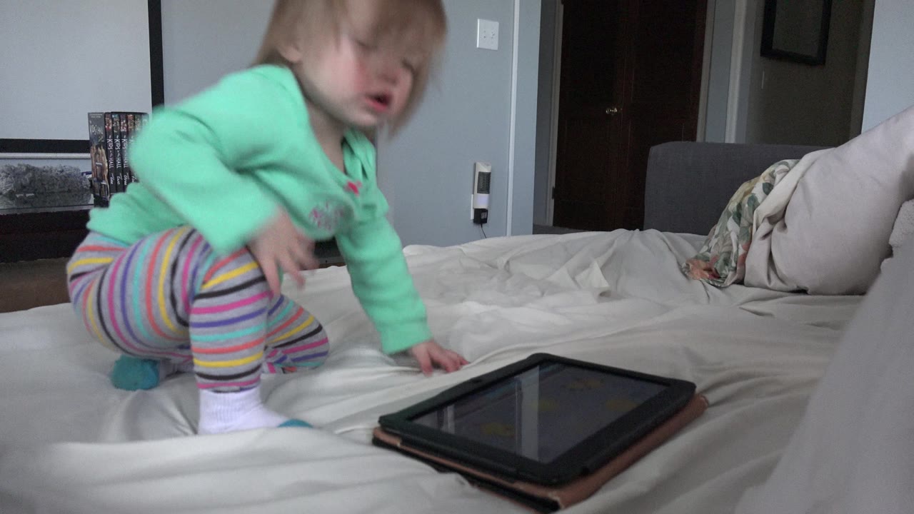 The child leans farther across the white bed toward the tablet, with the gray bedroom wall and dark door visible behind her.