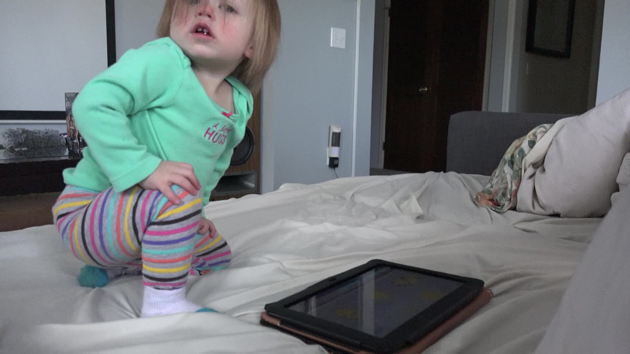 The toddler sits more upright on the bed beside the tablet, looking upward while pillows and a doorway sit in the bedroom background.