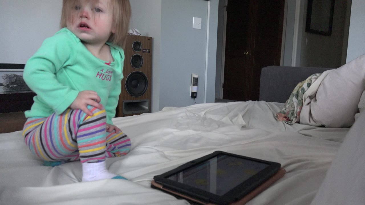 Young child sitting on a bed near a tablet in a home interior.
