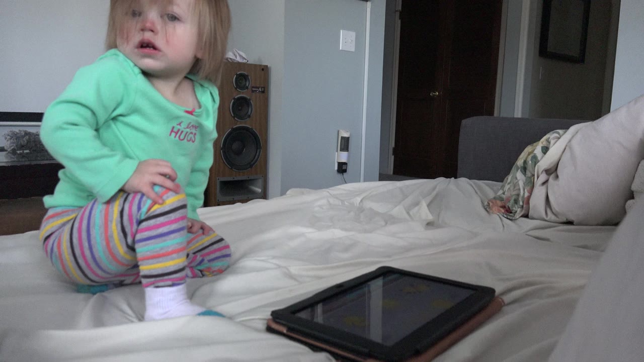 The child remains seated on the bed in the Westminster home, facing slightly toward the camera while the tablet rests in the lower foreground.