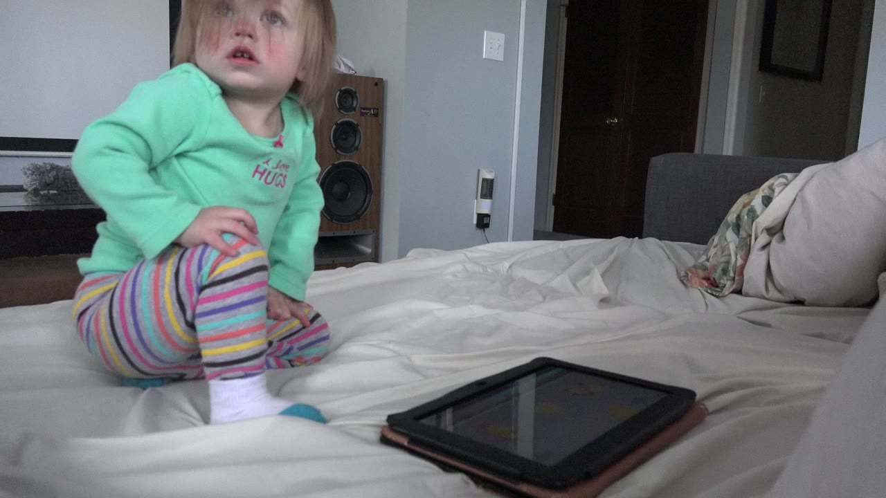 A young child in Westminster, Colorado sits on a white bed beside a dark tablet, with pillows and a speaker visible in the bedroom.