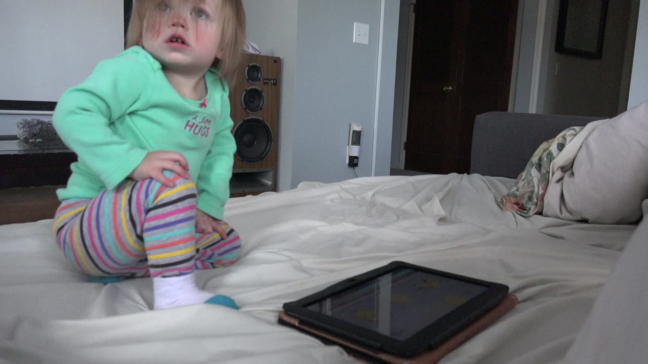The child remains seated on the bed in Westminster, Colorado, looking upward while the tablet lies closer to the camera on the bedding.