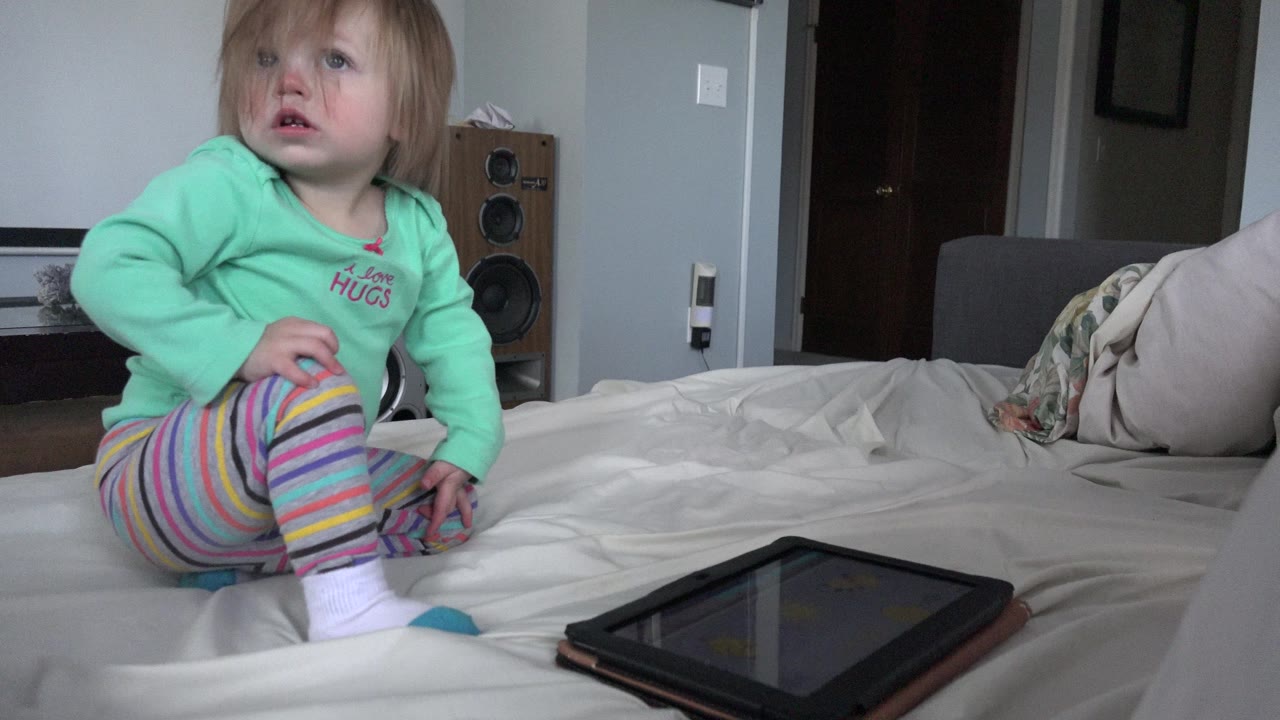 A young child sits upright on a white bed near a tablet in a Westminster, Colorado home interior, with pillows and a speaker visible behind them.