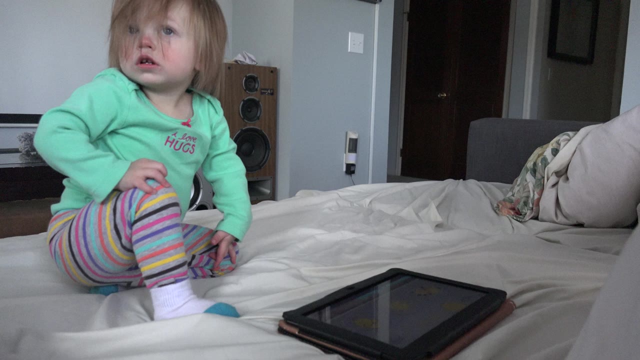 The child remains seated on the bed in the Westminster home, looking upward while the tablet lies in the lower foreground on the bedding.