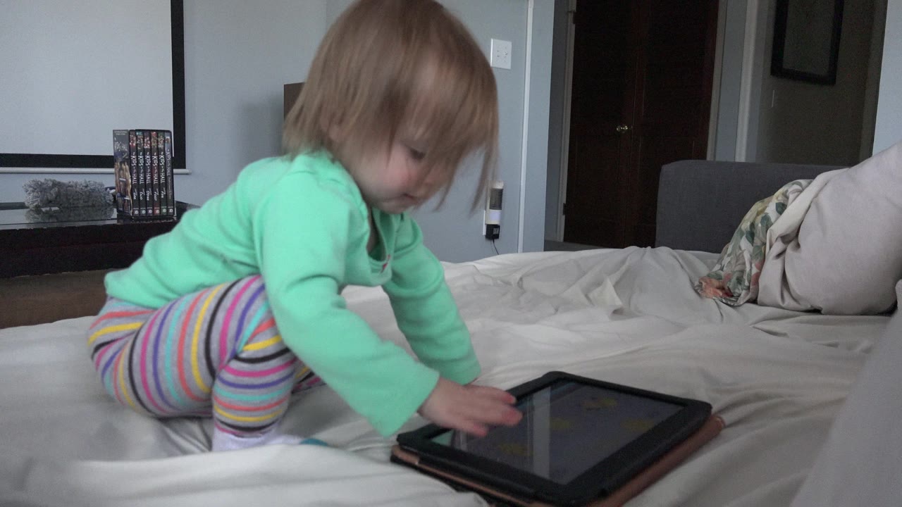 In Westminster, Colorado, a blond baby in a mint green shirt kneels on a white bed and reaches both hands toward a tablet near the lower edge of the frame.