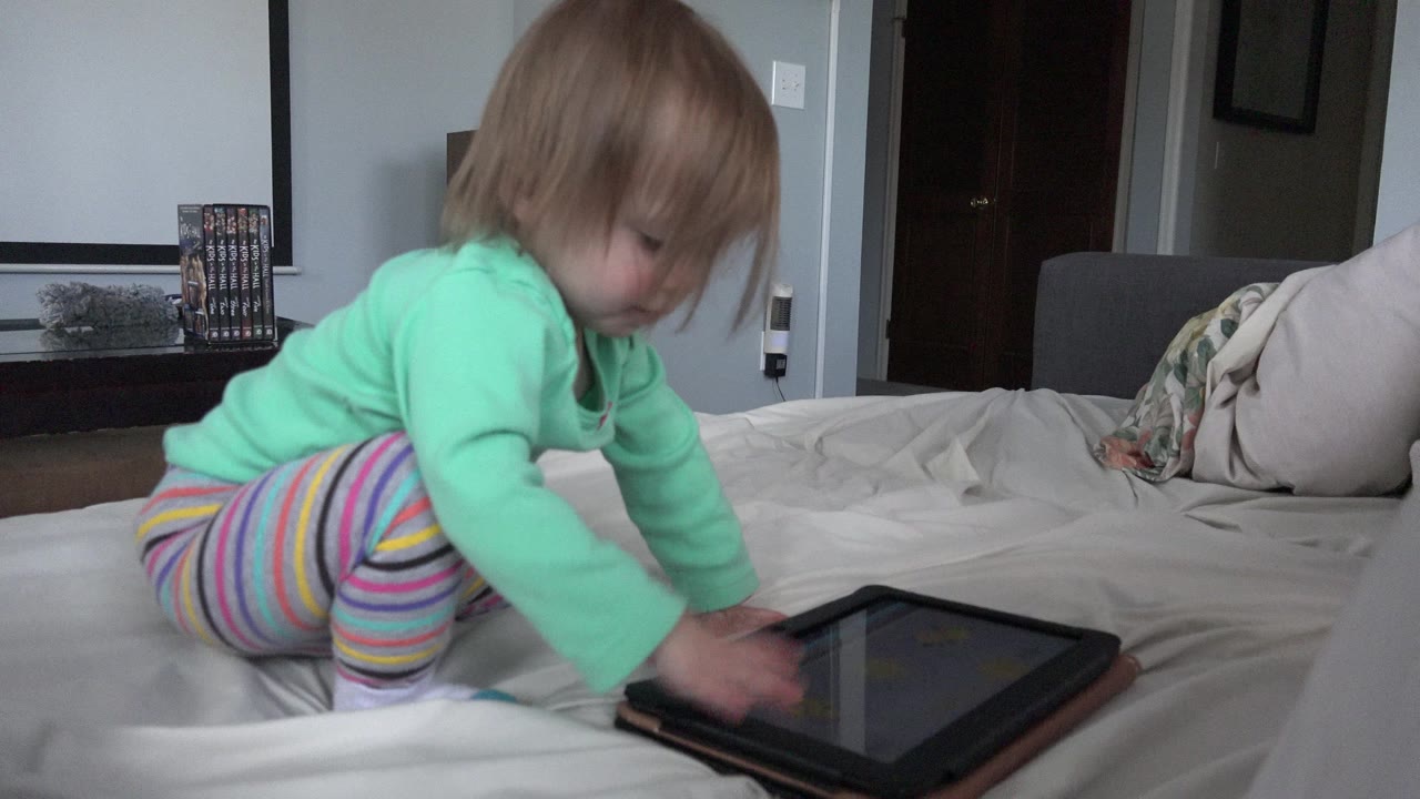 The baby remains kneeling on the bed in Westminster, Colorado, leaning forward as one hand hovers over the tablet screen and the other steadies near the case.