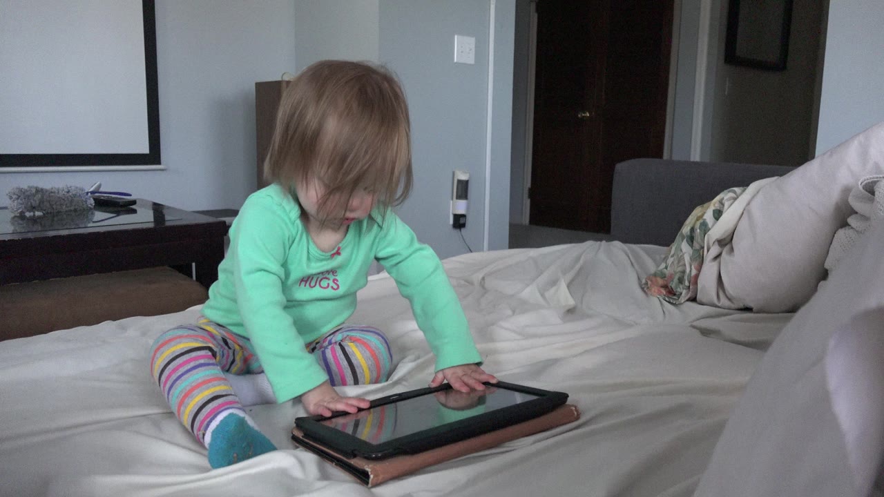 At 0:00 in Westminster, Colorado, a young child in a green shirt sits cross-legged on a white bed and touches a tablet near the lower edge of the frame.