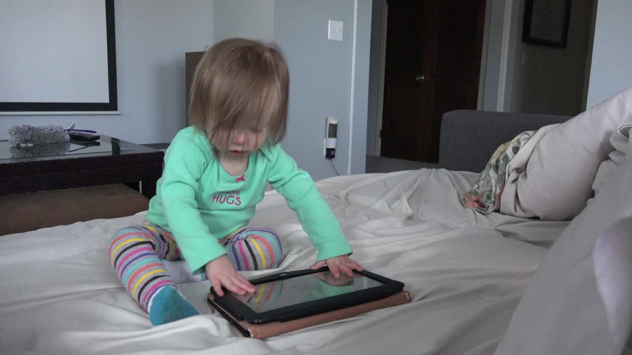 Toddler sitting on a bed and touching a tablet.