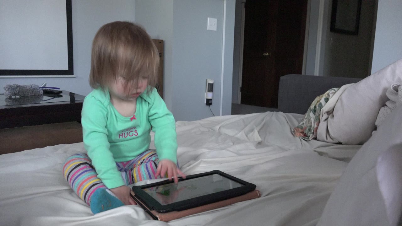 Toddler sitting on a bed and touching a tablet.