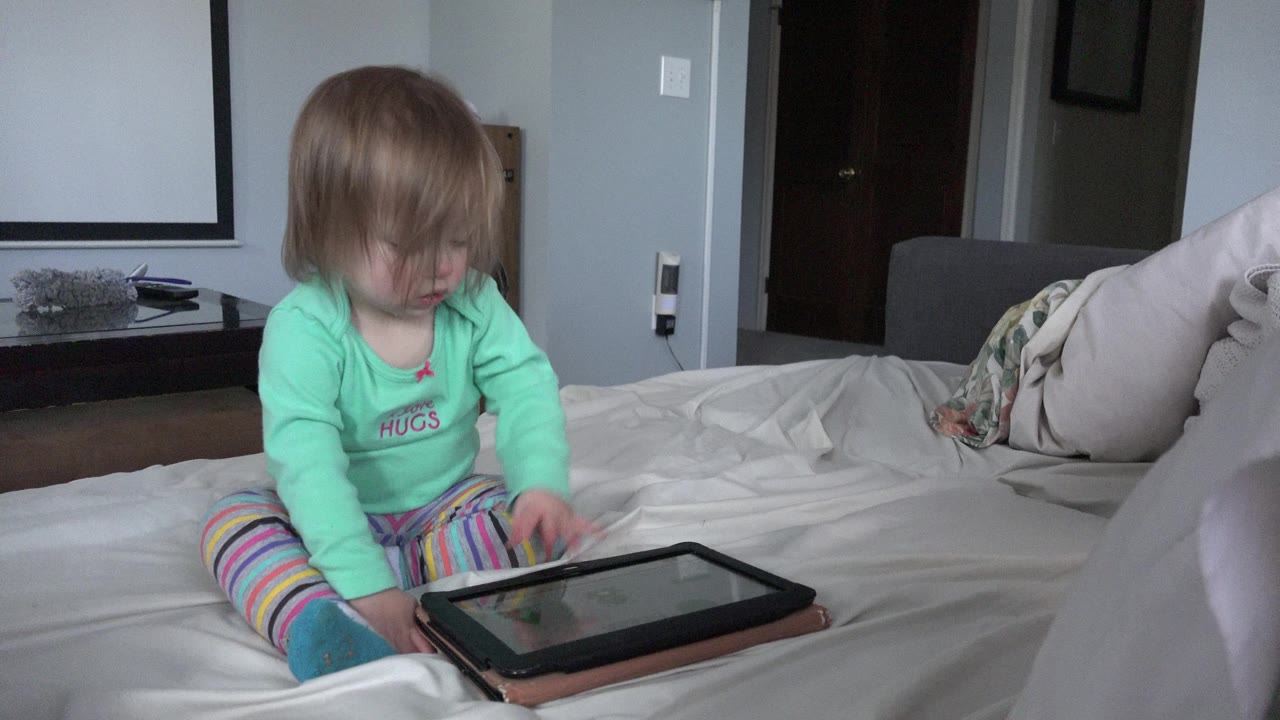 The toddler remains seated on the bed in the Westminster, Colorado home interior, with one hand near the tablet screen and pillows visible behind her.