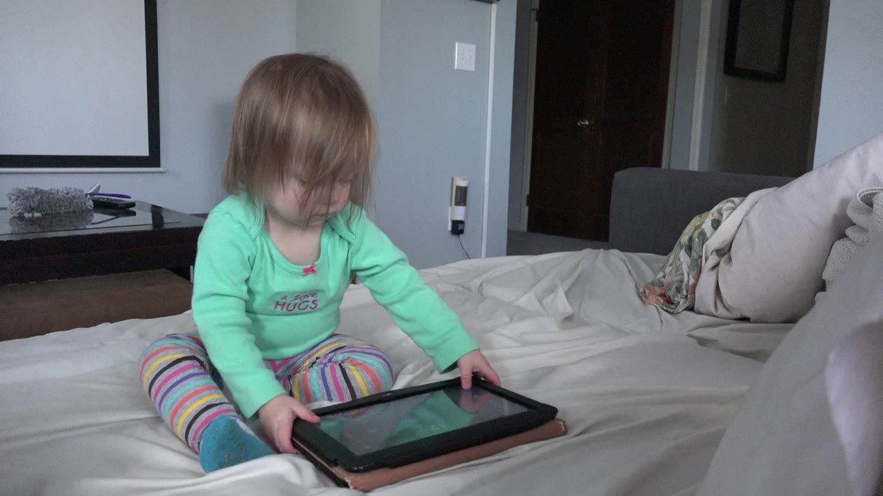 Toddler sits on a bed holding a tablet.