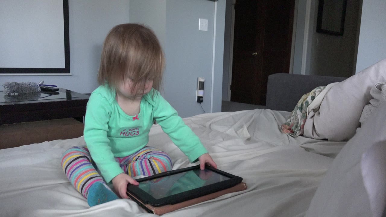 The child remains centered on the bed in Westminster, Colorado, with both hands near the tablet and her hair partly covering her face.