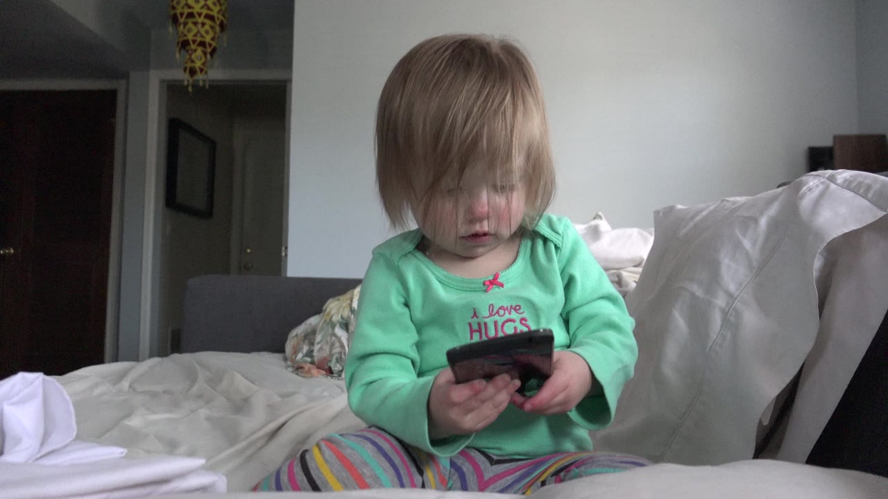 In Westminster, Colorado, the baby remains seated on the bed and looks down at the cellphone, with rumpled bedding and a dark doorway in the background.