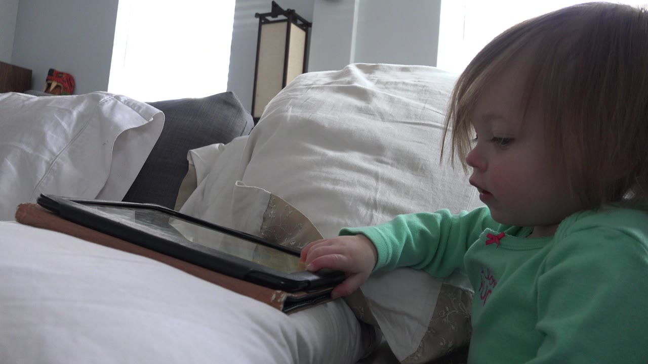 Young child reaches toward a tablet on white bedding.