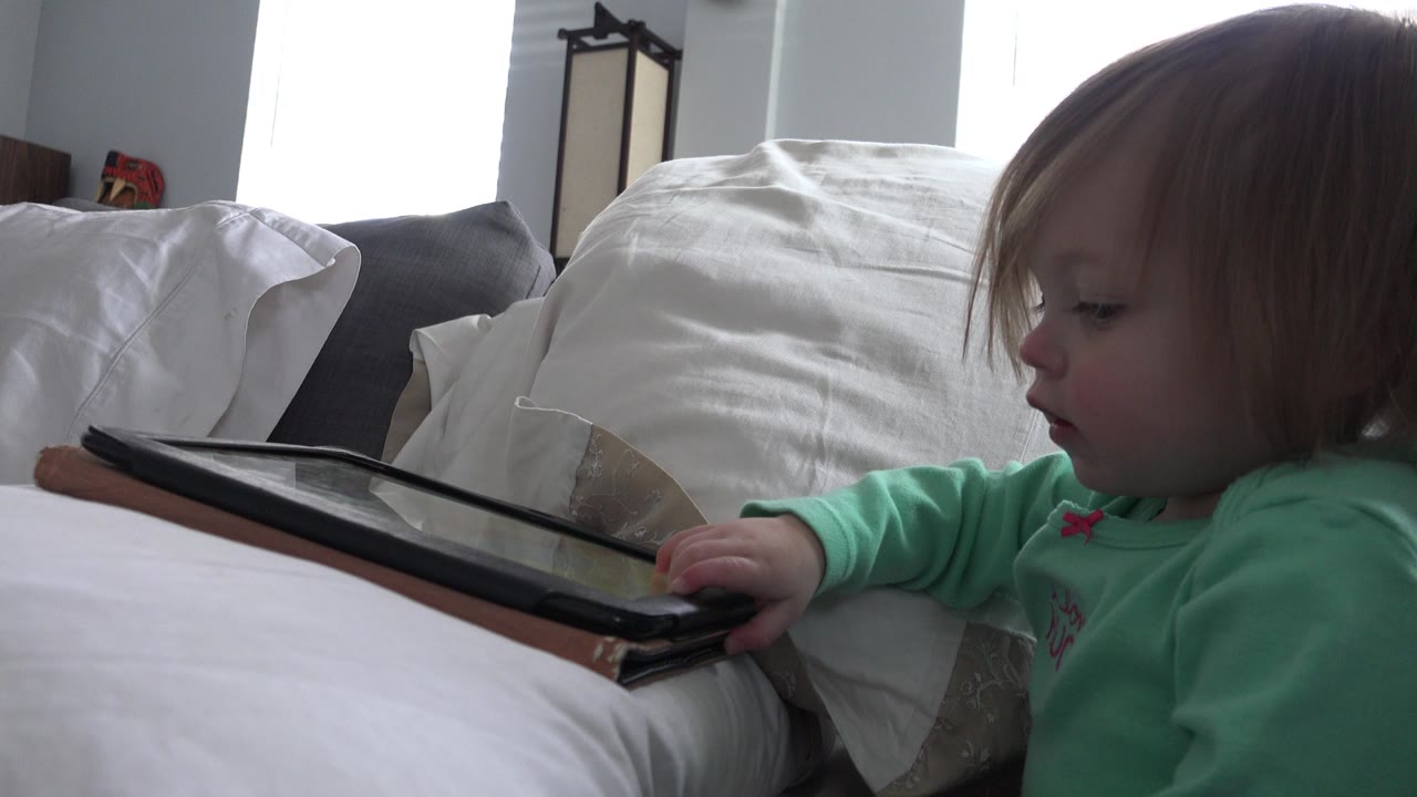 At 0:01 in Westminster, Colorado, the child stays in profile with one hand on the tablet case and white bedding filling the foreground.