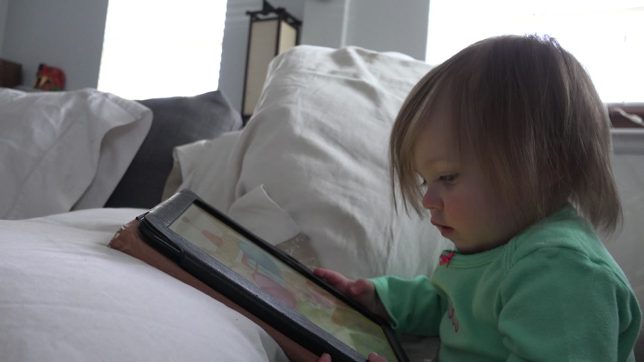 At 0:00, a young child in a mint green top sits in profile on a white bed in a Westminster, Colorado home, looking down at a black-cased tablet.