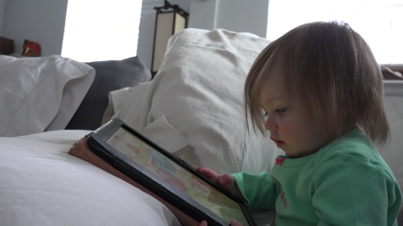 At 0:01, the child remains seated on the white bed, holding the tablet close while daylight from the rear windows outlines the room.