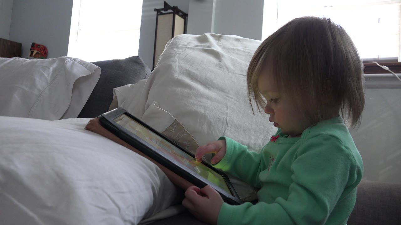 Blond child in green clothing holding a tablet on a bed