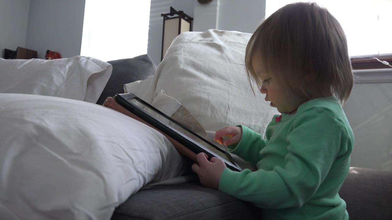 At 0:00, a young child in a green top sits in profile in a Westminster, Colorado home, holding a tablet near white pillows and a bright window.