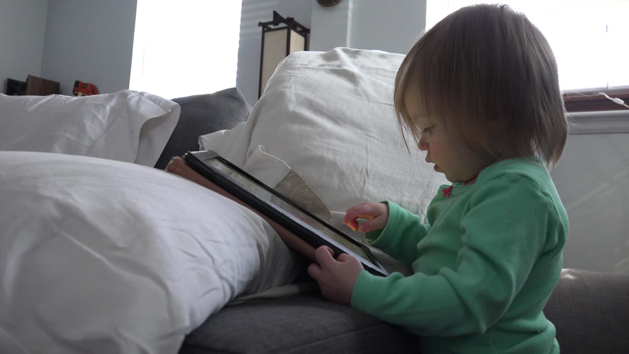 At 0:01, the child looks down toward the tablet screen while seated next to a large white pillow in the Westminster, Colorado interior.