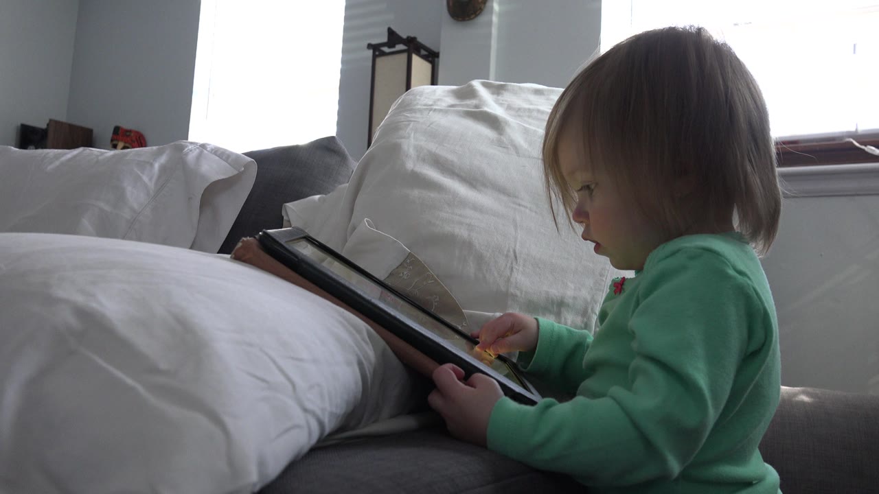 Young child seated beside white pillows while holding a tablet.