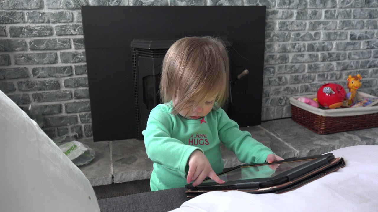 At 0:00, a young child in mint green pajamas leans over a black tablet in a Westminster, Colorado home interior, with a gray brick fireplace and toy basket behind.
