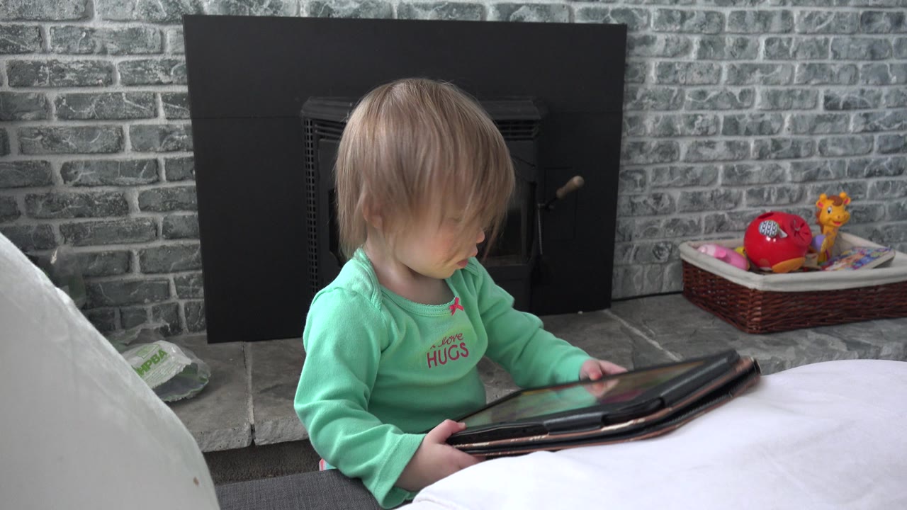 The child remains near the white couch in Westminster, Colorado, looking down at the tablet while the black stove and basket of toys sit in the background.