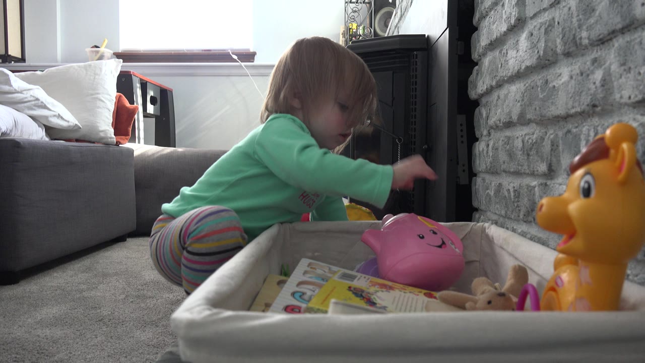 A toddler girl in Westminster, Colorado reaches toward toys in a fabric box while sitting low on the carpet, with the couch, window, stone wall, and dark stove behind her.
