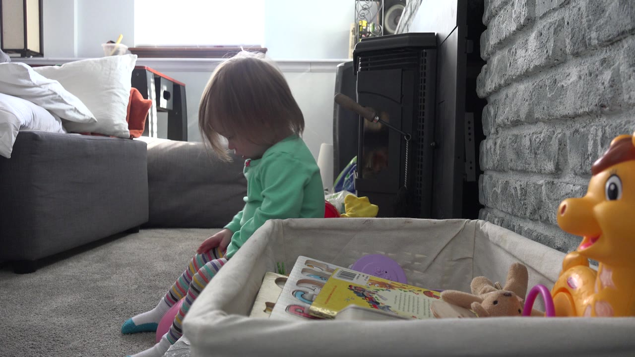 Young child sitting on a carpeted floor beside a toy bin in a living room.