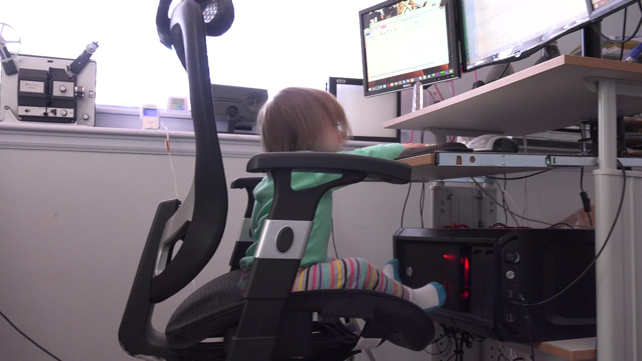 At 0:01, the girl remains seated sideways in the office chair, with striped pants visible and the desktop tower glowing beneath the desk.
