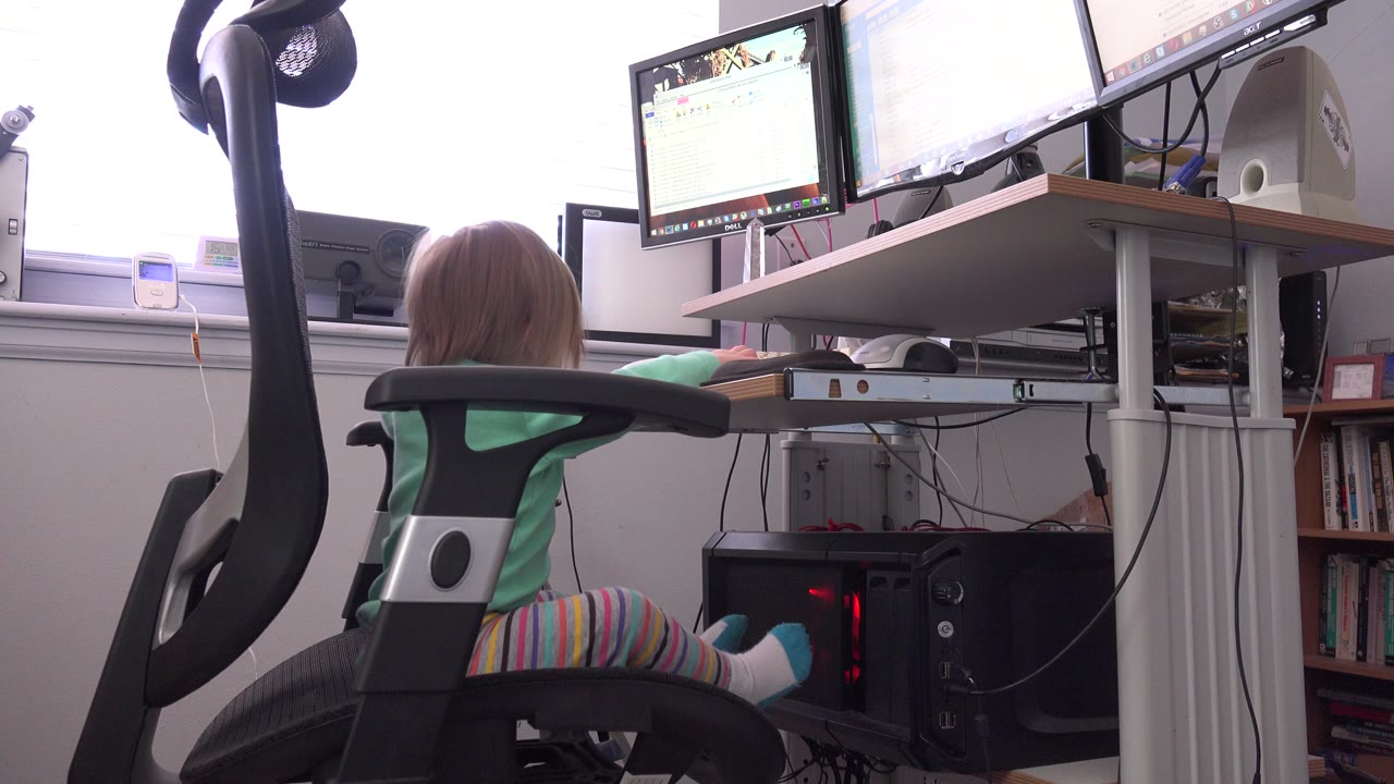At 0:01, the child remains seated at the workstation, with one arm extended toward the desk and bright window light behind the monitors.