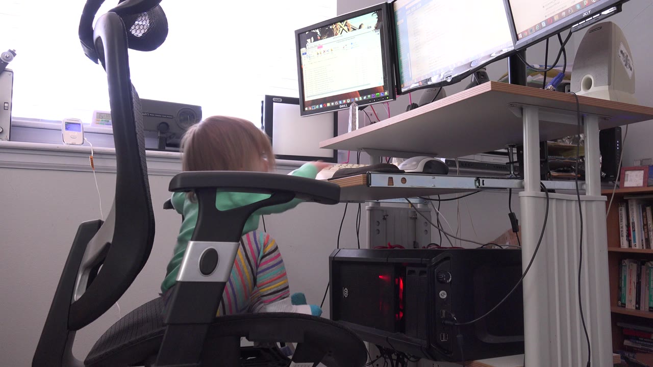 Small child seated in an office chair beside a computer desk.