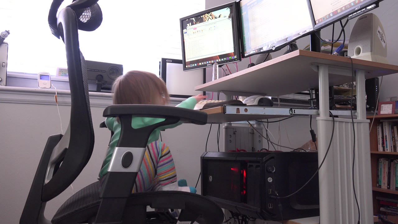 The child remains seated in the black office chair, facing the computer desk with two monitors, a keyboard, mouse, speaker, and daylight from the window.