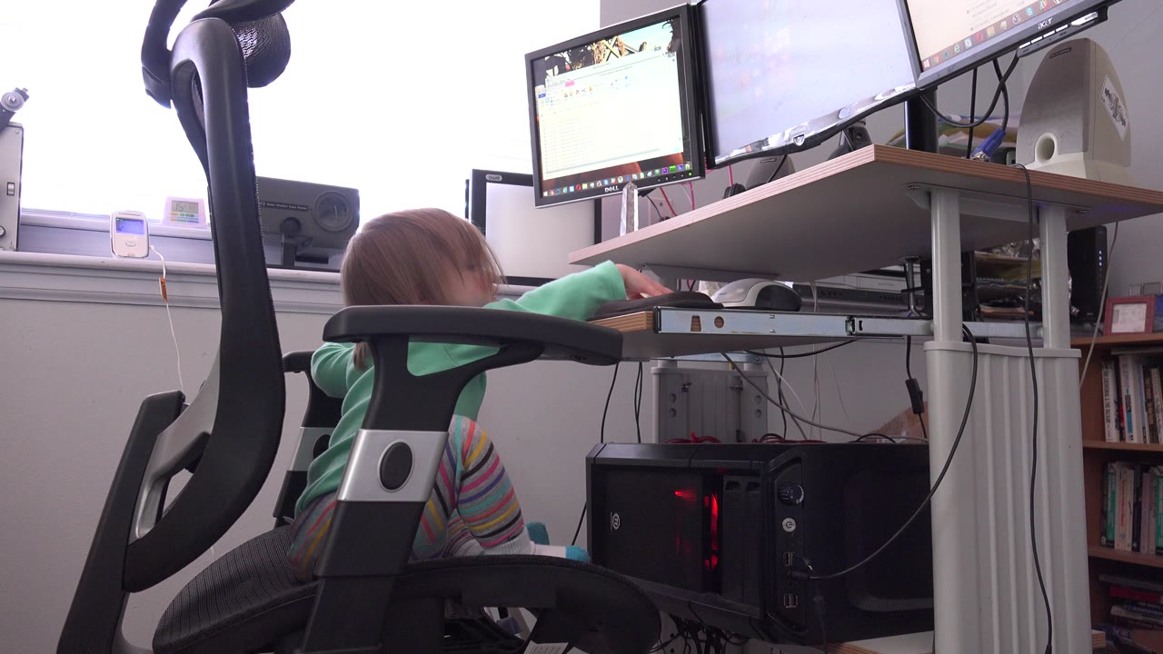 At 0:01 in Westminster, Colorado, the child stays low behind the chair arm while facing the desk, with the monitor screens, mouse, tower, cables, and window light visible.