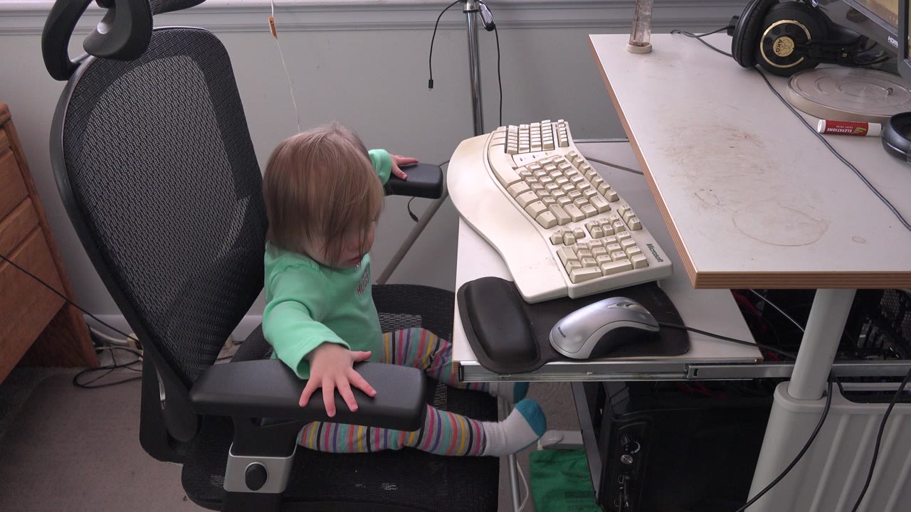 Baby girl sitting on a black office chair beside a computer keyboard.