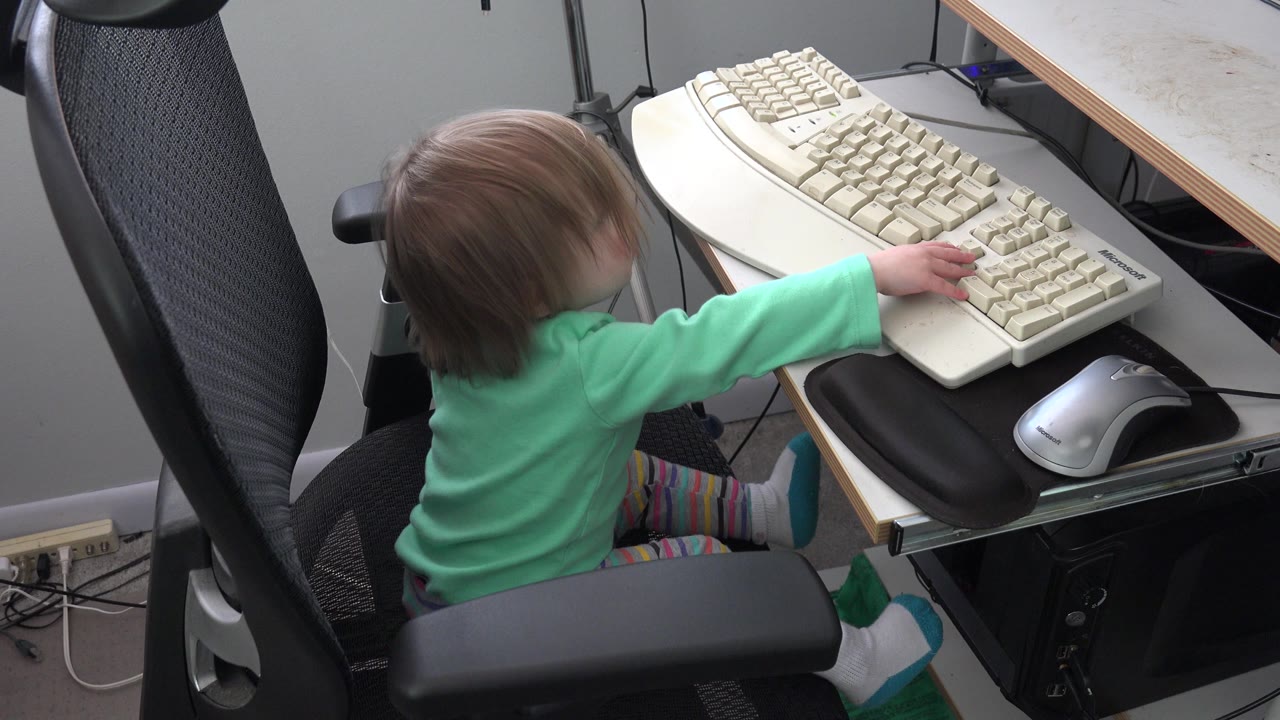 At 0:01 in Westminster, Colorado, the child leans farther over the keyboard tray, with the silver mouse and desk edge clearly visible beside the beige keyboard.