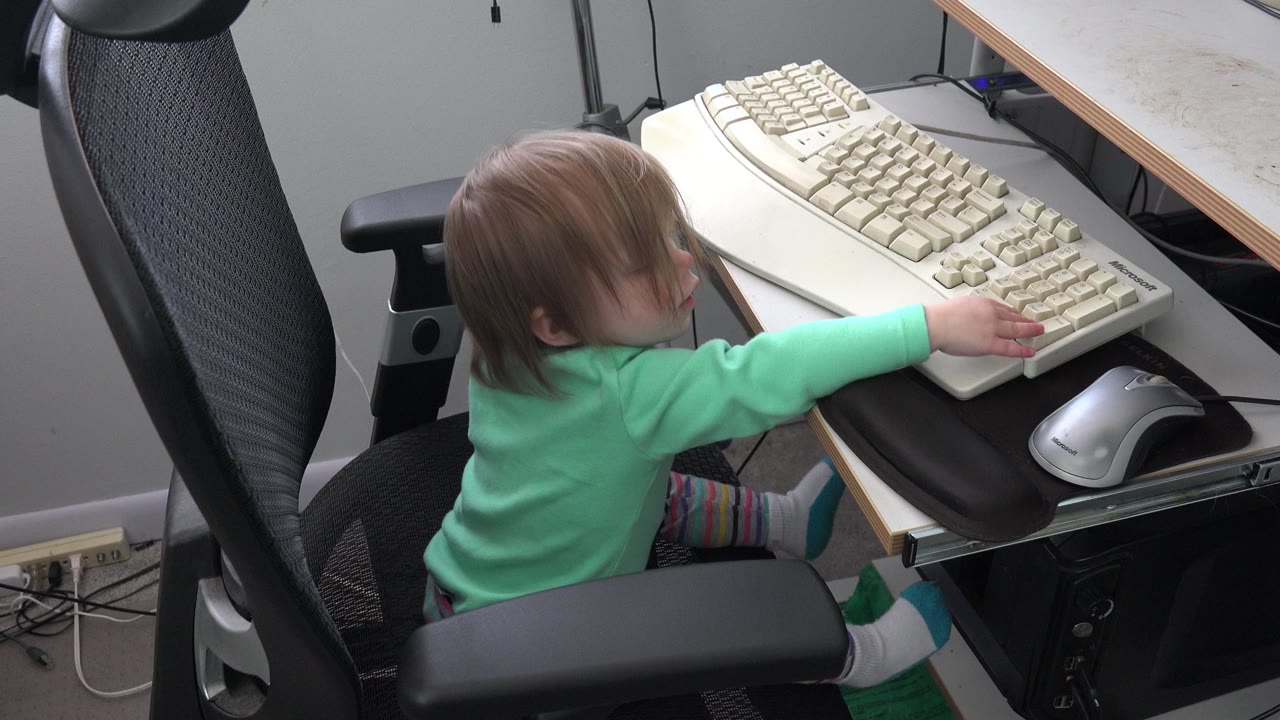 Child in a black office chair reaching toward a beige computer keyboard.