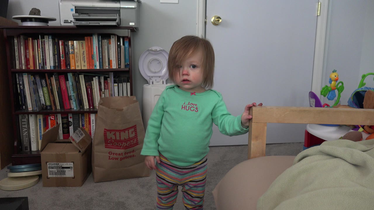 Toddler girl stands beside a bed frame in a bedroom.