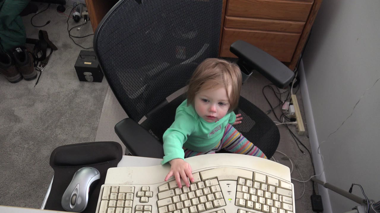 Toddler seated at a desk reaching toward a computer keyboard.