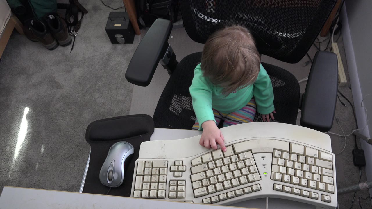 At 0:00 in Westminster, Colorado, a young child in a mint green shirt sits at a desk and points toward the center of a large ergonomic keyboard.